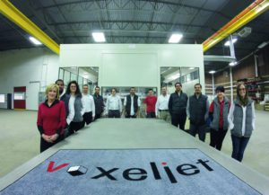 Das Team von Voxeljet America steht zum Größenvergleich an der 4000 mm langen Bauplattform - im Hintergrund die VX4000.
